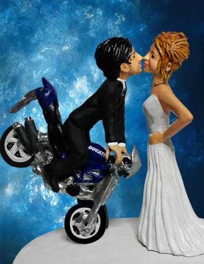 Cake topper sposi di terracotta raffiguranti la sposa in piedi mentre bacia lo sposo che è sulla moto davanti a lei.